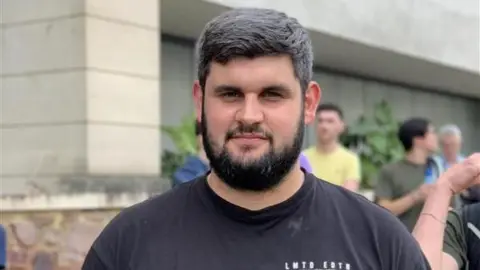 A man stood outside a hospital that has been evacuated. He is wearing a black t-shirt. 