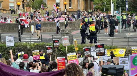 BBC Two large groups of protesters, separated from one another by a road and lots of police officers. One group is holding England flags and union jacks, while the other side holds placards reading 'refugees welcome'.