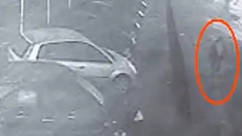 Merseyside Police A grainy still from a CCTV clip showing a man with his hood raised walking past a car show room. 