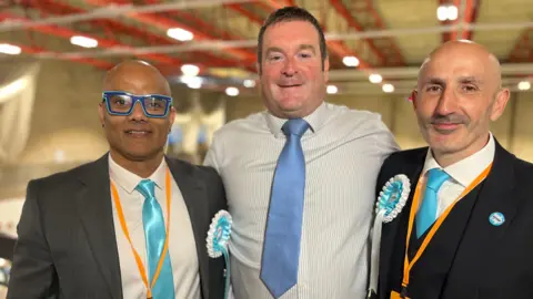 Karan Maheshwari, Ryan Coogan and Howard Tobias are all standing side by side. Maheshwari is wearing blue glasses, a white shirt, blue tie and grey blazer. He is wearing a Reform UK rosette. Coogan has short brown hair and is wearing a blue and white stripped T-shirt and blue tie. Tobias is wearing a white shirt, blue tie and black suit jacket. He is also wearing a Reform UK rosette and sticker.