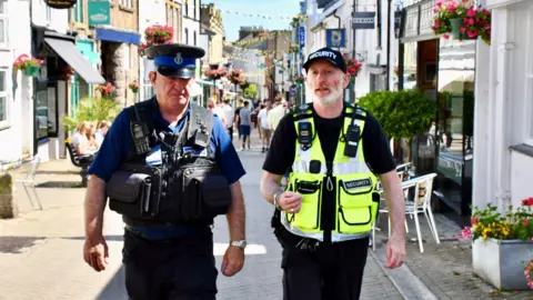 Devon and Cornwall Police A police community support officer and a security guard walking down a road in Penzance on a sunny day.