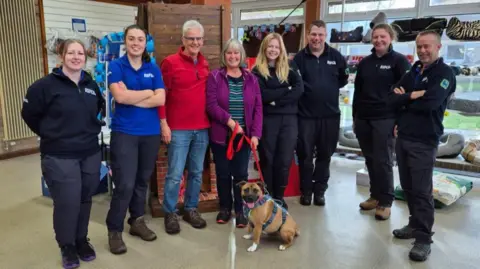 RSPCA Honey a small brown dog with white paws. She has a red lead and is surrounded by her new owners and the RSPCA team.