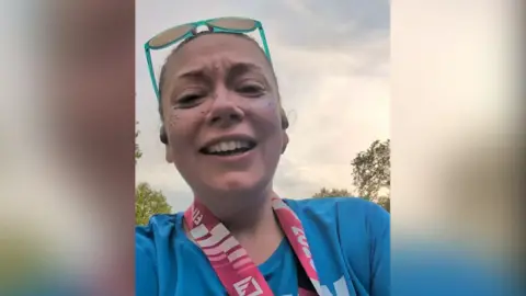 Micky Marsden with brown hair tied back and green sunglasses resting on her head wearing a blue t-shirt with the PAC charity logo and the words Positive Action in the Community. She is at the finishing line of the London Marathon with the medal around her neck. She is smiling.