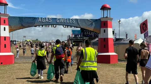 BBC People walking around the Boardmasters festival site. Some are wearing yellow high-vis jackets and are picking up litter.
