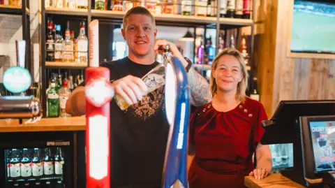 Marston's A man with short hair on the left is wearing a black top and standing next to a woman with brown and blonde hair who is wearing red. They are standing behind the bar and bottles are on shelves in the background.