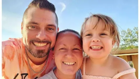 Tracey Watkins pictured smiling with blue sky behind her and her partner to her left and daughter to her right