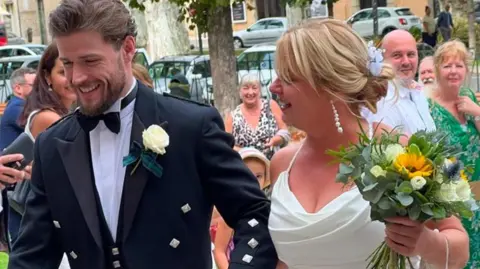 Instagram Callum Kerr, a dark haired man wearing a kilt, smart jacket and a bow tie with a white rose as a button hole. He is holding the hand of an older woman, his mother, who has blonde hair and is wearing a wedding dress. She is holding a bouquet of flowers. Both are smiling.