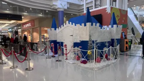 A blue and white castle style grotto with a small fence around it, and shops away to the left of the attraction