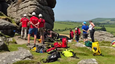 Bovey Tracey Fire Station A group of people wearing helmets gather beside large rocky outcrops on a hillside, with rescue equipment spread out on the grass and rolling countryside in the background.