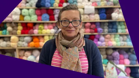 A woman wearing glasses, striped pink top and blue cardigan stands in front of a shop display of balls of wool.