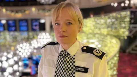 BBC/Gem O'Reilly Cdr Clair Kelland in a nightclub. She is looking at the camera with a neutral expression and is in uniform: a white shirt, shoulder lapels, name badge and checkered scarf known as dicing. Her hair is blond