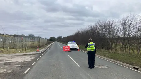 A police officer in high visibility jacket with her back to the camera stands in the middle of the road in front of a police car with its lights on and a red sign saying road closed