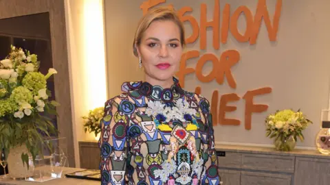 David M. Benett/Dave Benett/Getty Images Bianka Hellmich is wearing a very colourful and busy high-necked dress, and there is a 'Fashion for Relief' sign on the wall behind her. She has blonde hair pulled back, and bright pink lipstick.