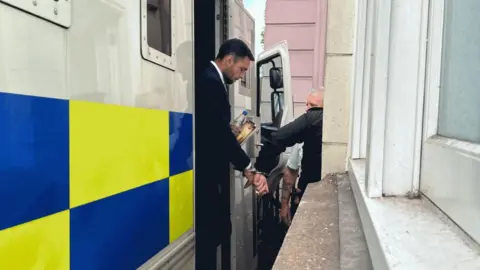 BBC Dylan Pounds being taken out of a police van outside court. He is handcuffed to a prison officer. He is wearing a suit and white shirt and is carrying a bottle of water and a book. 