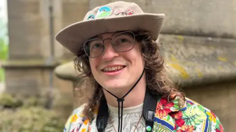 A man in a beige floppy hat and colourful shirt smiles to the camera. He wears glasses and has a black strap dangling around his neck.