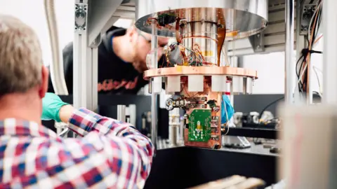 Oxford Ionics A green computer chip attached to a large copper coloured fixture with two men, both wearing gloves, working on it