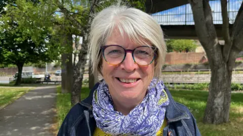 A woman with short grey hair, smiling, wearing glasses, a purple and white scarf and a denim jacket standing in front of an out-of-focus footbridge and road with vehicles in the distance.
