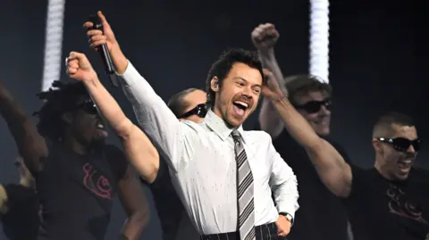 Harry Styles performs at the Brit Awards. He is wearing a grey office shirt and tie combination.