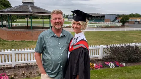 Andy Beasley A red haired man in a green open collared shirt, stood next to him is his daughter in a graduation cap and gown with a police uniform underneath. they are both smiling at the camera.