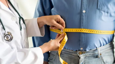 A person wearing a blue shirt and denim jeans is having their waist measured using a yellow tape. The person doing the measuring is wearing a white coat and has a piece of medical apparatus around their neck, it is black and silver. 