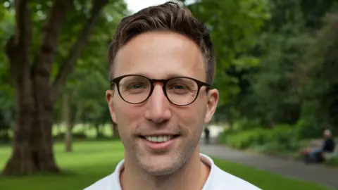 Matt Cortland Matt Cortland, a man with short, light brown hair, smiling at the camera while standing in a park. He is wearing dark-rimmed glasses and a white polo shirt. In the background there are tall, mature trees and a man sitting on a park bench. 
