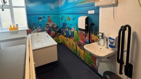 Northamptonshire Health Charity A ward bathroom which has a window and a bath on the left and a basin on the far right. The end wall and part of the wall on the right is decorated with a colourful mural which shows the sea, a beach, fish, dolphin, animals snorkelling, a submarine and plants. 