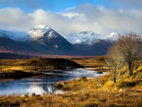 Greg Crawford Snow-capped mountains rise majestically under a partly cloudy sky, their rugged slopes casting deep shadows. In the foreground, a calm reflective loch winds through golden-brown moorland dotted with sparse trees.
