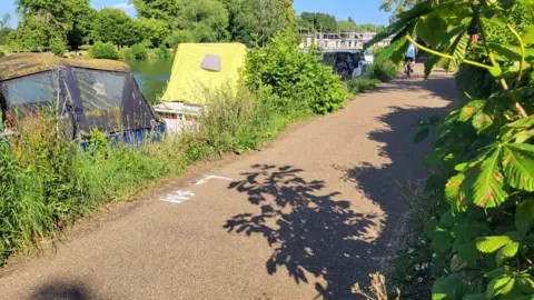 The Thames towpath near Folly Bridge on a sunny day. There are boats moored by the towpath. The bridge could be seen in the distance.