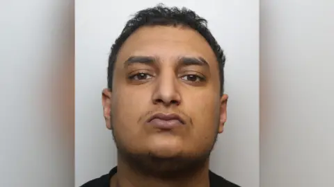 A close up crop of a mugshot. Janjua has brown eyes, brown hair, a thin moustache and thin beard.