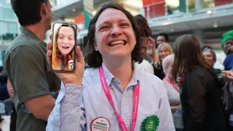 Green Party supporters congratulate candidate Florence Cullen-Davies - who is at home about to give birth and appears live via WhatsApp video on a mobile phone - after she is declared a winner in the 2026 London Borough of Hackney