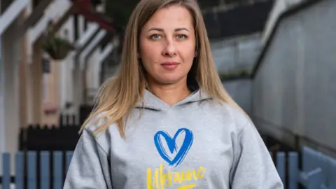 FSWC A woman with long fair hair looks into the camera. She is wearing a grey hoodie with a blue heart and 'Ukraine' written in yellow.