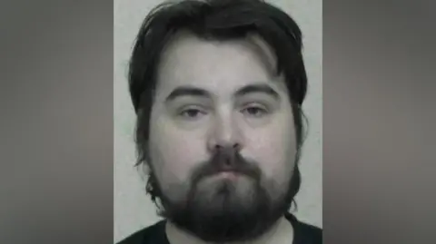 Mugshot of McLean. He has large head with very pale skin and scruffy long black hair and beard.