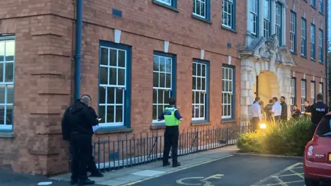 At least half a dozen people are standing outside an entrance to the building, which is on the right of the photo. Three other people are in the middle and left of the image.