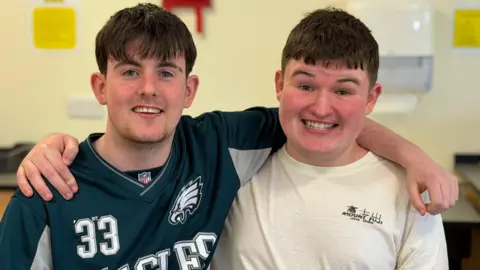 Two boys hug each other and smile for the camera at the life skills classroom. They both have short black hair with a fringe.