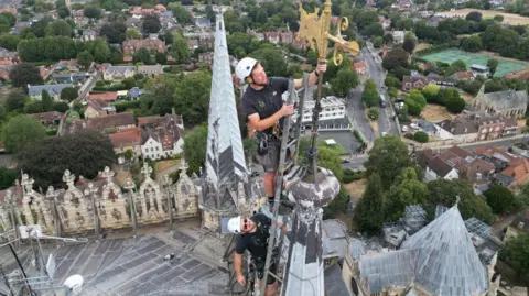 Two men in black tops and shorts, in harnesses and with white helmets are standing at the top of a ladder looking at a golden weather metal vane at the top of a cathedral's pinnacle. Below them is a city with buildings and trees.