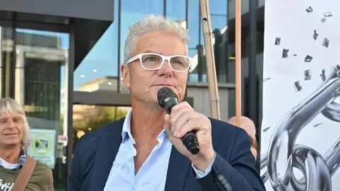 A man wearing white-rimmed glasses and a dark suit, holds a microphone