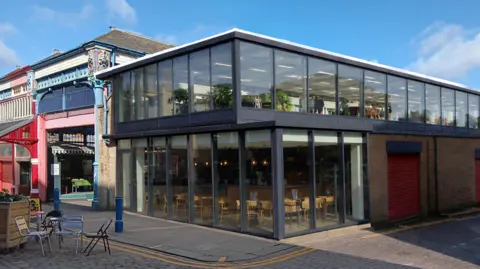 Andrew Wilson/ Same Skies Think Tank Image shows the exterior of a two-floored glass building with chairs and tables inside attached to a traditional market hall.