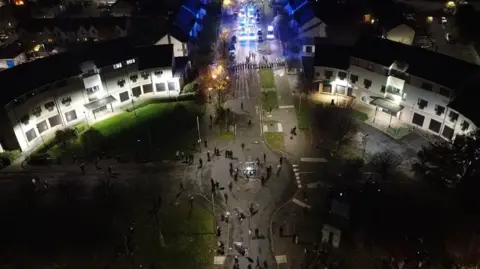 PA Media A drone shot of police forming a barricade while a group of people stand at the other end of a road. It is dark and they are lit by streetlights. 