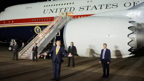 Reuters Donald Trump is standing in front of a large white aircraft. He is wearing a blue suit and a yellow tie. Men dressed in suits are standing behind him.