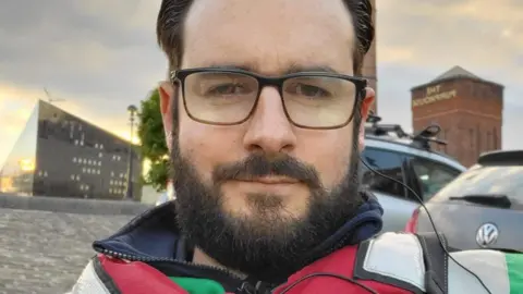 Handout Lewis Bridson, who has black hair gelled back and a black beard, poses for a selfie in a red life-jacket with a view of Liverpool's waterfront in the background.
