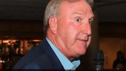 A side view of a man who has grey hair and is wearing a dark blue suit jacket and a light blue shirt. 