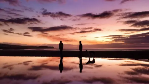 Two people and a dog are silhouetted on a beach at sunset.