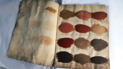 Guzelian/ Lorne Campbell A close up picture of a dye ledger which has twelve swatches of different dyes, including reds and browns, you can faintly see dates written below each of them.