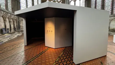 A large white box inside a huge medieval chapter house with stained glass windows and decorative tiled floor. There is an entrance and exit to the box, which makes it appear dark inside, where people can walk into to see the document.