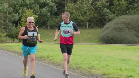 Debbie running in a park wearing a vest that reads "Stop Glioblastoma". Graeme is running wearing a vest that reads "The Brain Tumour Charity".