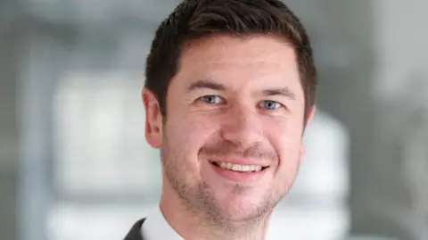 Jordan Buchanan is a man with dark hair. he is wearing a white shirt under a black jacket. He is smiling.