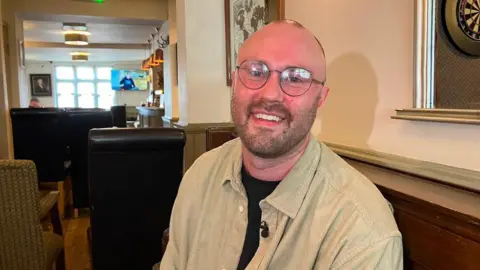 A bald man wearing glass is sitting indoors in a pub. He has a black t-shirt on with a beige shirt over the top of it. He is smiling for the photograph.
