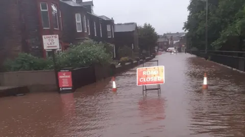 Flooding at Station Road, Wigton. There is a row of two-storey houses to the left. On the right is a black metal fence with tree behind it. Between the road is completely flooded with dark brown water. A red "road closed" sign is in the middle of the flooded road with a traffic cone either side.