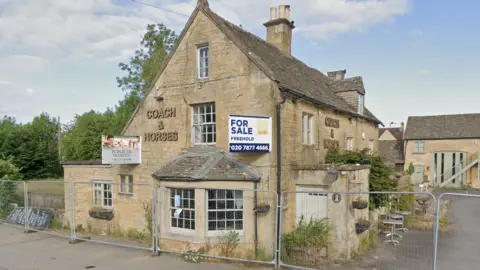 Google A closed pub in the Cotswolds, made of Cotswold stone. It has a large bay window on the ground floor and is surrounded by metal construction fencing. It has signs that read "for sale" and a metal sign that reads "Coach & Horses".