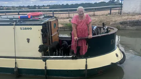 Kate Bradbrook/BBC A woman standing on a canal boat. She is smiling and has her long hair tied back. She is wearing a red dress. The boat is on water with fields behind her. 